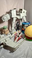 Shows large white AT-AT Walker with Darth Vader figure, plush Baby Yoda doll, X-Wing Fighter and other ships, Star Wars blanket, and Kenner Ewok Tree Hut