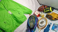 Photo showing entire lot including green waterproof jacket, two tennis rackets with case, tennis racket clock, Franklin baseball glove, two footballs, shuttlecock can, blue umbrella and poncho.