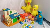 Wide view of the Sesame Street toy collection including Fisher Price Clubhouse, Big Bird plush dolls, Cookie Monster bank, and various small toys and rattle