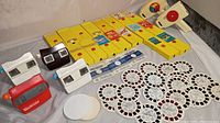 Photo showing 4 View Master viewers in white, black, and red, 15 yellow Fisher Price cartridges, and multiple round View Master reels