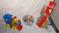Shows the Mr. Machine windup robot on the right, the blue and yellow windup train on the left, and two transparent plastic see-thru clock toys in the center.