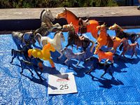Photo showing 14 vintage plastic horse figurines in various colors and poses on a blue tarp outside with wood surface background.