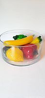 Large clear glass bowl holding five colorful glass fruit pieces: yellow banana, yellow lemon, red bell pepper, green bell pepper, and dark purple eggplant, all with gold windings.