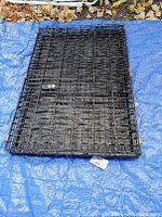 Folded black metal dog crate showing flat rectangular base and mesh wire construction on blue tarp with some rust on the latch.