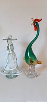 Image showing two handblown glass figurines side by side on a flat surface, a colorful green and red rooster with a clear base and a clear glass woman figurine wearing a large hat.