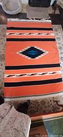 Full view photo of the large vintage hand-knotted wool rug showing the bright orange base with black and white stripes and central diamond motif in blue and black.