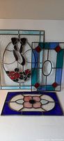 Three stained glass window panels with one panel featuring a dogwood flower with visible cracked blue piece, another geometric panel with blue and green diamond accents, and a third panel with a central pink flower framed by blue border.
