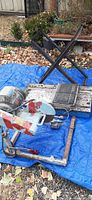 View of the full tile saw setup including saw machine, metal stand, blue tarp beneath, and some tools and accessories around.