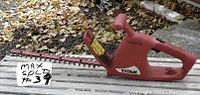 Side profile view of the red Titan electric hedge trimmer placed on a wooden surface with fallen leaves in the background. The Titan logo is visible on the body. Shows the blade length and handle design.
