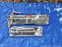 Two manual rail tile cutters placed side by side on a blue tarp outdoors, showing overall condition and size comparison.