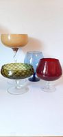 All four handblown glass items arranged together on white background: beige compote, blue snifter, red snifter, green textured compote bowl.