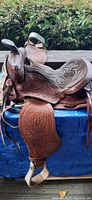Side view of the brown tooled leather western saddle showing detailed floral tooling, metal stirrup with leather covering, and leather straps. Saddle sits on blue surface with green plants in background.