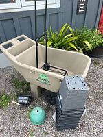 Overview of beige Ames Easy Roller Jr. wheelbarrow with black luggage cart inside, green ceramic pot, grey plastic pots stacked and black outdoor post light beside it on gravel.
