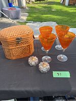 Photo of four heavy orange textured cocktail glasses, two small glass pumpkins, and woven basket chest on black surface outdoors.