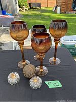 Full view of four amber glass tealight holders, two small glass pumpkins, and decorative ball on black surface outdoors