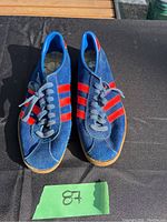 Pair of blue suede Adidas Gazelle shoes with red stripes, gum sole, and white laces pictured on black cloth surface with green lot number card