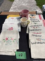 Full view of vintage embroidered dish towels with days of the week and two Disney mugs outside in sunlight.
