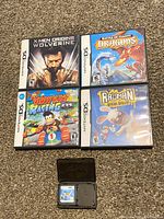 Five Nintendo DS game cases placed on carpet, showing front covers for X-Men Origins Wolverine, Battle of Giants Dragons, Diddy Kong Racing DS, Rayman Raving Rabbids, plus Tetris Party Deluxe cartridge in black case.