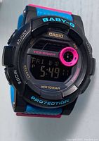 Front view of Casio Baby-G digital watch showing tide graph, day/date/time display, BABY-G branding in blue, and colorful bezel details