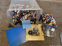 Wide shot showing a clear plastic tote filled with mixed Lego bricks and parts, two Lego baseplates (one large blue, one small light blue) placed in front, and several small plastic bags with assorted Lego pieces including wheels and gears