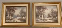 Two framed handpainted winter landscape artworks side by side on the wall in matching gold-tone frames with cream and gold matting showing a river, birch trees, and a cabin.