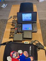Black Nintendo DS open with both screens visible, top screen damaged, showing calendar menu. Two charger adapters and two carrying cases visible, including a Mario softness case.