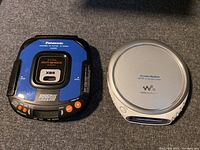 Panasonic Shockwave blue portable CD player and silver Sony Walkman CD player side by side on gray surface.