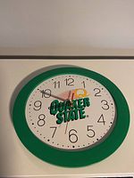 Front view of the clock showing green frame, white clock face with black numbers, Quaker State logo, and clock hands at about 11:50.