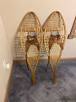 Pair of vintage wooden snow shoes with webbing and leather binding shown upright against a wall.