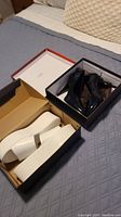 Two pairs of shoes, one black pair with 6-inch heels and one white platform sandal pair, shown in their boxes on a bed.