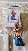 Full view of the ornament positioned beside its original box showing the merman figure holding watersports gear with blue glitter tail.