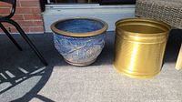Two large planters placed side by side outdoors on a patio floor, showing the brass/gold metal planter and the blue clay planter with gold trim and grape motif.