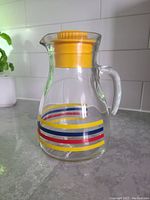 Clear glass pitcher with yellow, blue, and red stripes, yellow stopper lid on top, placed on a countertop with a white tiled backsplash.