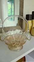 Pressed glass fruit bowl with detailed fruit motif, arched clear handle, scalloped rim, and elevated base on a white shelf with a mirror and decorative vases in the background.