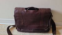Full front view of brown leather Samsonite shoulder bag with flap and shoulder strap, showing overall condition and size relative to wall and floor.