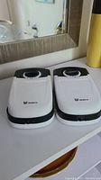 Two battery-operated pet food feeders with white lids and black bases sitting closed side-by-side on a white surface.