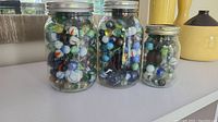 Three clear glass jars with metal lids filled with vintage marbles of various colors and sizes on a white surface.