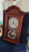 Full front view of the cherry stain wooden pendulum wall clock showing the arched top, clock face, and pendulum behind glass.