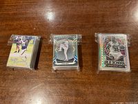 Three sealed stacks of sports trading cards on a wooden surface, each stack individually shrink-wrapped or sealed in plastic, showing football, baseball, and basketball cards.