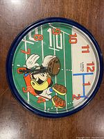 Front view of round wall clock showing Mickey Mouse holding a football on a green football field background with orange numbers.