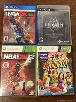 Four video game cases displayed containing NBA 2K15 for PS4, Skyrim Legendary Edition for PS3, NBA 2K12 for Xbox 360, and Kinect Adventures for Xbox 360.