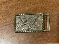 Front view of the brass belt buckle showing the detailed soaring eagle and mountain background with rope border.