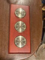 Full view of the wooden weather board with three metal instruments in a vertical row on a wooden table.