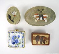 Top view of four trinket boxes showing different materials and designs: porcelain floral, wooden bear inlay, and two stone boxes with butterfly and flower inlays.