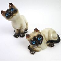 Pair of vintage ceramic Siamese cat figures with bright blue eyes, one sitting and one lying down, shown from front and top angles.
