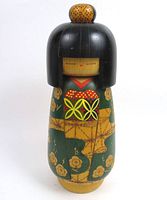 Front view of the Kokeshi doll showing painted facial features, hair bun, and green kimono with floral and geometric decorations.