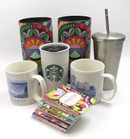 Two architectural design ceramic mugs featuring colorful motifs, one white Starbucks ceramic travel mug with green logo, one stainless steel tumbler with metal straw and lid, and a large set of colorful Starbucks Reserve tasting cards spread out.