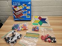 Full view of 10 game set components including game boards, playing pieces in bags, and colored sticks on a wooden table with box in background.