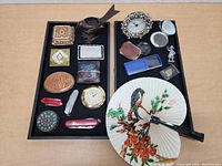 Two trays featuring assorted buckles, small collectible items, pocket knives, a hand fan, clock face, mirrors, and figurine.