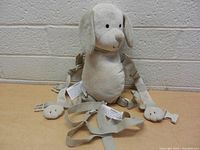 Front view of beige plush dog child safety harness backpack with straps and buckles on a beige surface in front of a light gray brick wall